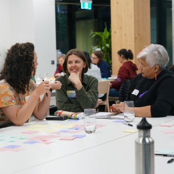 Three women in conversation at a table covered with coloured paper pieces during a workshop session.